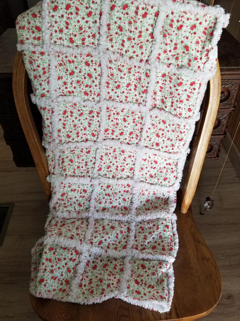 Pu&ograve; includere: Una copertina per neonati di colore bianco e rosso con stampa floreale e un bordo bianco, soffice e sfilacciato. La copertina &egrave; piegata e appoggiata su una sedia di legno.
