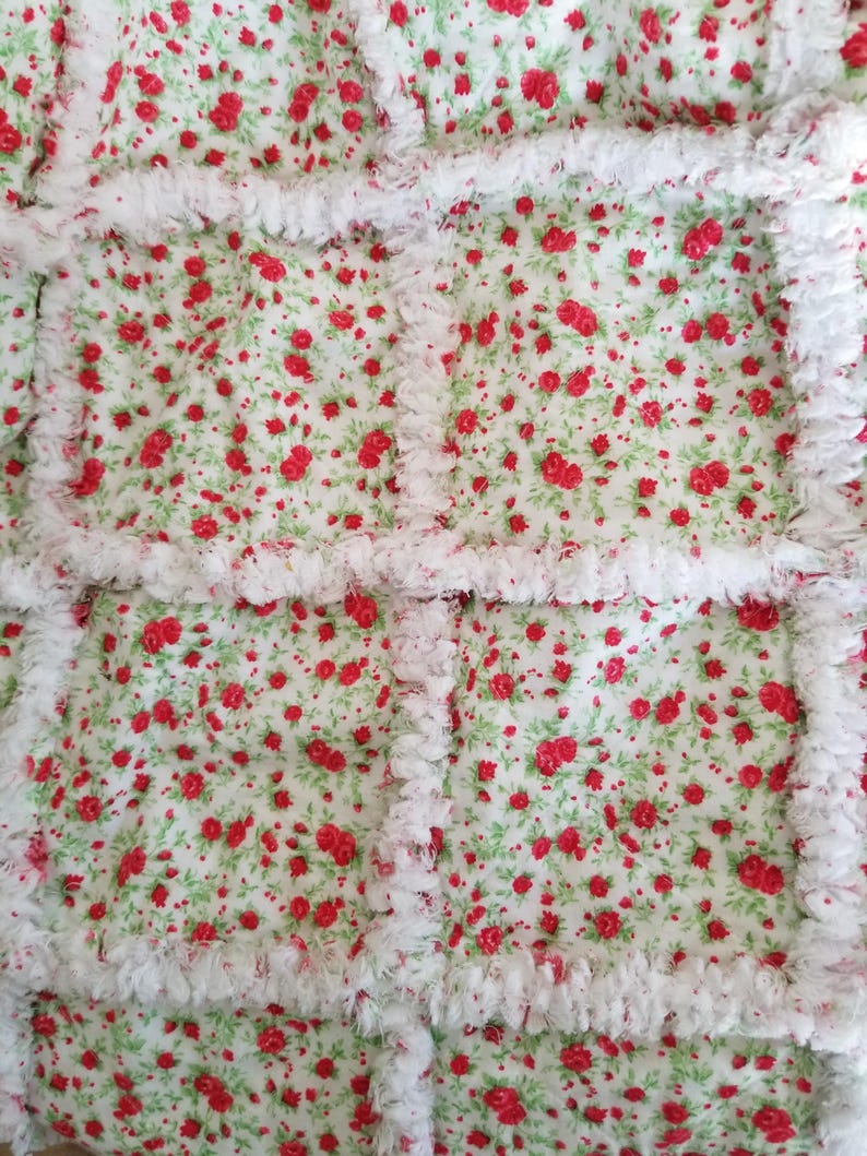 Pu&ograve; includere: Una trapunta floreale bianca e rossa con un bordo bianco, soffice e sfrangiato. La trapunta &egrave; realizzata con quadrati di tessuto con un motivo ripetitivo di piccole rose rosse su sfondo bianco.