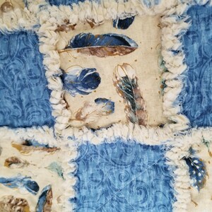 K&ouml;nnte beinhalten: Eine blaue und cremefarbene Patchwork-Decke mit ausgefransten Kanten. Die Decke besteht aus verschiedenen Stoffen mit einem Federmuster und einem blauen Paisley-Muster.