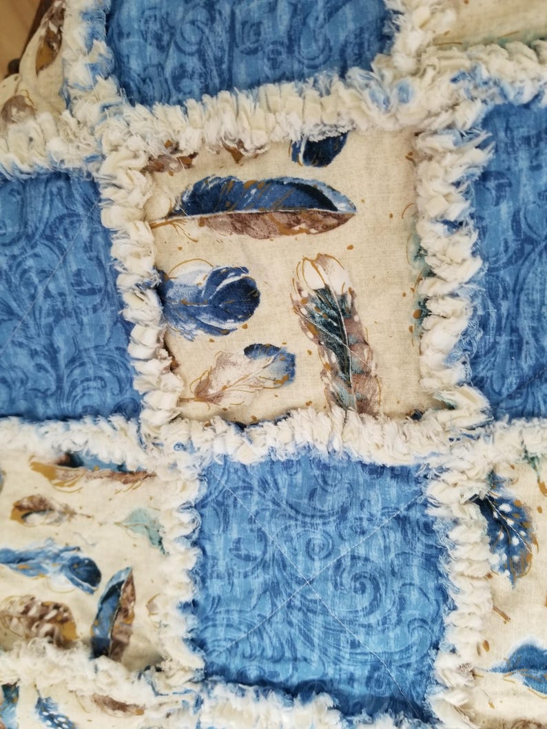 K&ouml;nnte beinhalten: Eine blaue und cremefarbene Patchwork-Decke mit ausgefransten Kanten. Die Decke besteht aus verschiedenen Stoffen mit einem Federmuster und einem blauen Paisley-Muster.