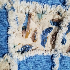 K&ouml;nnte beinhalten: Eine Nahaufnahme einer blauen und cremefarbenen Patchwork-Decke mit ausgefransten R&auml;ndern. Die Decke besteht aus verschiedenen Stoffen mit verschiedenen Mustern, darunter ein cremefarbener Stoff mit einem braunen Federmuster.