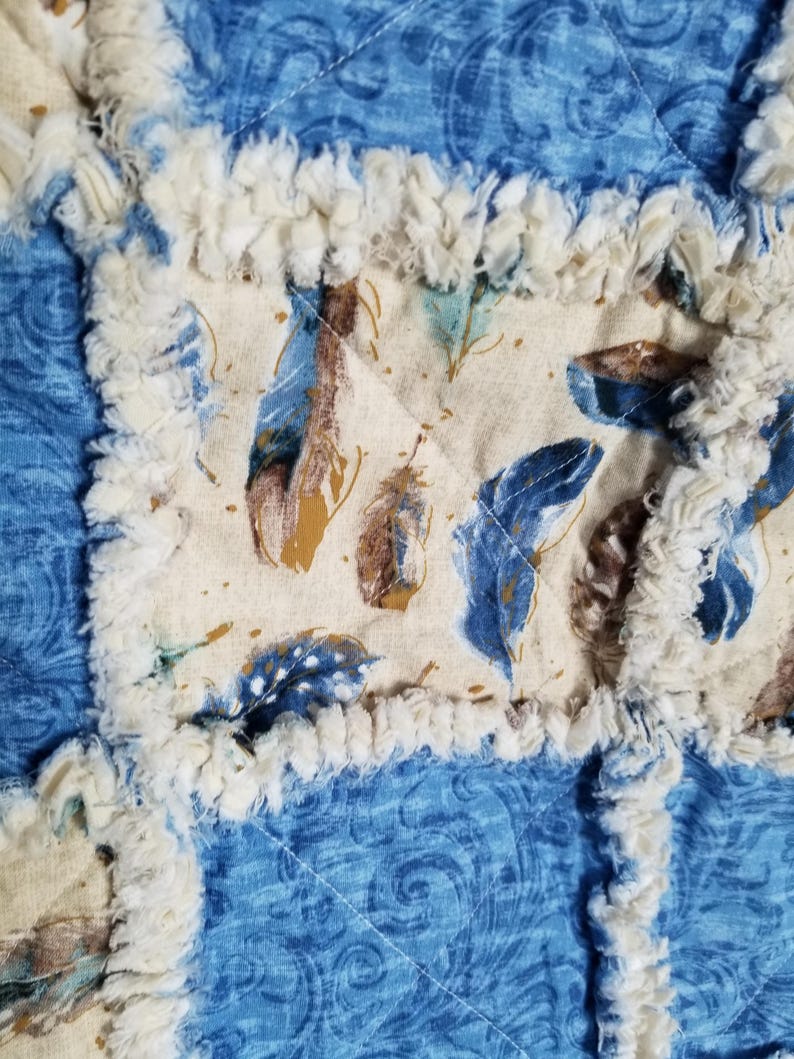 K&ouml;nnte beinhalten: Eine Nahaufnahme einer blauen und cremefarbenen Patchwork-Decke mit ausgefransten R&auml;ndern. Die Decke besteht aus verschiedenen Stoffen mit verschiedenen Mustern, darunter ein cremefarbener Stoff mit einem braunen Federmuster.