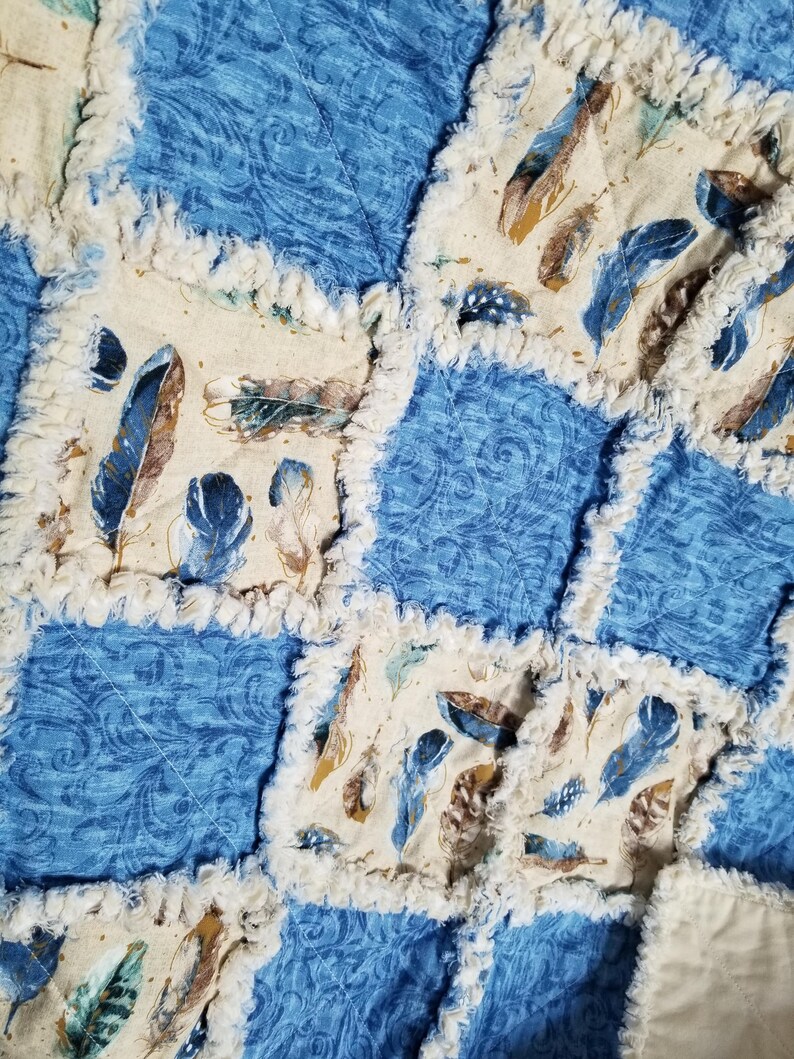 K&ouml;nnte beinhalten: Eine Patchwork-Decke in Blau- und Cremet&ouml;nen. Die Decke besteht aus Quadraten mit verschiedenen Mustern, darunter ein Federmuster. Die Decke hat einen ausgefransten Rand.