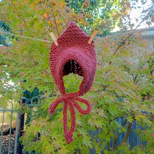 May include: A knitted burgundy bonnet with a pointed crown and a face opening, suspended by wooden pegs on a washing line. The bonnet features a tie closure and is set against a backdrop of green and yellow foliage.