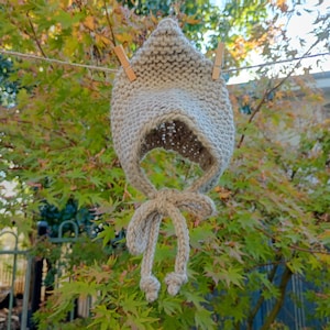 Pode incluir: Um gorro bege de malha com uma parte superior pontiaguda e uma abertura para o rosto. O gorro é suspenso por pregadores de madeira num varal. O gorro tem laços longos que são amarrados nas extremidades. O fundo apresenta folhagem verde e amarela.