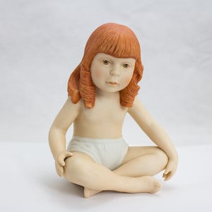 Puede incluir: Una figurita de porcelana de una niña con el pelo rojo sentada con las piernas cruzadas. Lleva un pañal blanco y tiene una expresión seria en la cara.