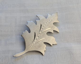 Könnte beinhalten: Silberfarbene Brosche in Blattform mit strukturierter Oberfläche. Die Brosche hat ein detailliertes Design mit spitzen Kanten und einem zentralen Adernmuster. Das Blatt ist leicht angewinkelt und zeigt seine komplizierte Handwerkskunst und elegante Form.