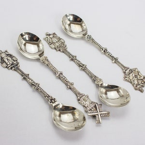 Puede incluir: Conjunto de cuatro cucharaditas de plata con mangos adornados con diseños de temática holandesa. Cada cuchara tiene un diseño diferente, incluyendo un molino de viento, una mujer con vestido tradicional, un hombre cargando cubos y un edificio.