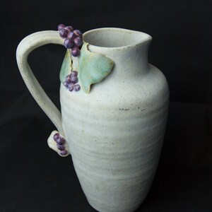 May include: A white ceramic pitcher with a textured surface and a handle. The pitcher is decorated with a grapevine design with green leaves and purple grapes.