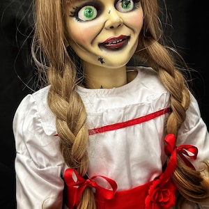 May include: A porcelain doll with long brown hair styled in braids with red ribbons. The doll is wearing a white dress with a red sash and a red rose. The doll has green eyes and a painted smile.
