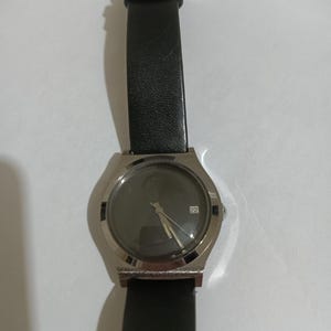 May include: A silver wristwatch with a black leather strap. The watch face is dark with silver hands and a date display. The strap is approximately 23 cm long.