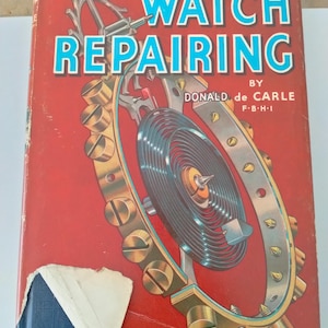 Op de afbeelding: Een vintage boek getiteld "PRACTICAL WATCH REPAIRING" door Donald de Carle. De rode omslag heeft blauwe en witte tekst en een gedetailleerde illustratie van horlogeonderdelen. De kaft van het boek is licht beschadigd.