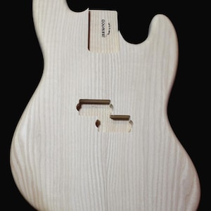 May include: Unfinished bass guitar body made of light wood with a grain pattern. The body has two cutouts for pickups and a label that reads "JBRWOOD Made in USA".