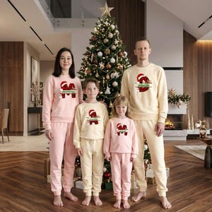 May include: A family of four wearing matching Christmas pajamas. The pajamas are light pink and cream-colored, with a Santa Claus graphic and the words "mommy" and "daddy" on the shirts. A Christmas tree is in the background.