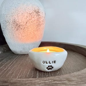 Puede incluir: Una vela encendida en un cuenco de cerámica blanca con el nombre "OLLIE" y una huella de pata. El cuenco tiene un interior amarillo y está sobre una bandeja de madera. Un jarrón blanco texturizado está en el fondo.