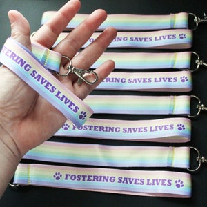 Könnte beinhalten: Ein Set aus sechs regenbogenfarbenen Lanyards mit dem Text "Fostering Saves Lives" und einem Pfotenabdruck. Die Lanyards sind aus weichem, gewebtem Stoff gefertigt und haben einen silbernen Metallverschluss.