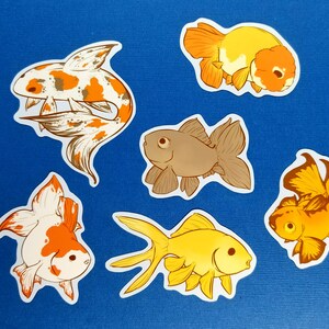 May include: Six goldfish stickers in various designs and colours, including orange, white, brown, and yellow, are arranged on a blue surface. A ruler is visible on the left side of the image, providing a size reference. The stickers are ideal for decorating.