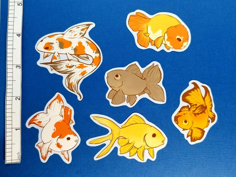 May include: Six goldfish stickers in various designs and colours, including orange, white, brown, and yellow, are arranged on a blue surface. A ruler is visible on the left side of the image, providing a size reference. The stickers are ideal for decorating.