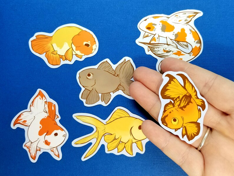 May include: A selection of goldfish stickers in different designs. The stickers depict detailed illustrations of goldfish in shades of orange, yellow, and brown, with white detailing. The stickers are on a white background.