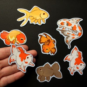 May include: A selection of goldfish stickers in different colours and designs. The stickers depict goldfish in shades of orange, white, brown, and gold. Some stickers have a koi-like pattern. The stickers are on a black background.