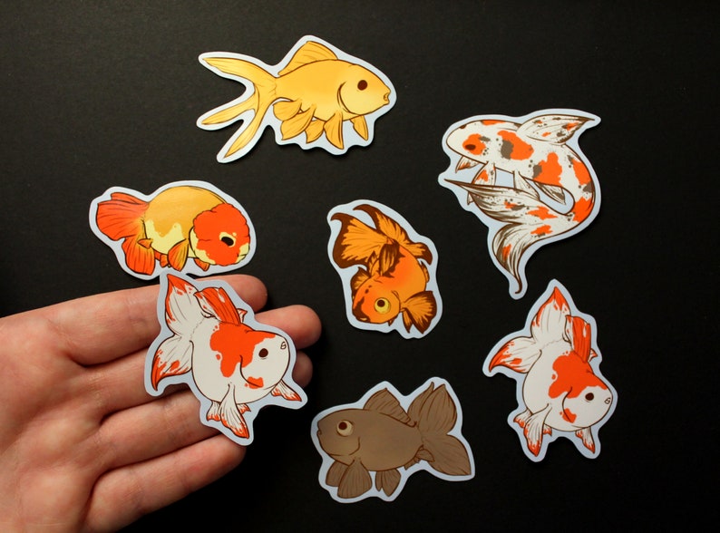 May include: A selection of goldfish stickers in different colours and designs. The stickers depict goldfish in shades of orange, white, brown, and gold. Some stickers have a koi-like pattern. The stickers are on a black background.