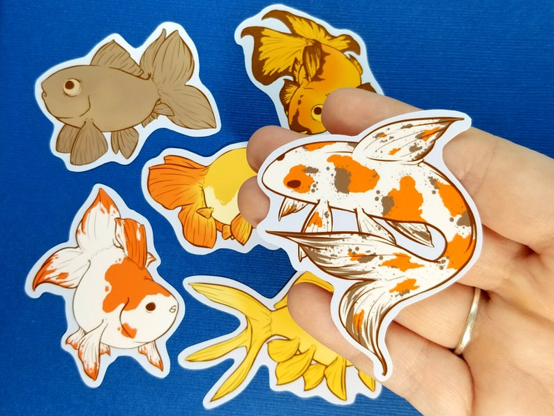 May include: A selection of goldfish stickers in different designs and colours. The stickers depict detailed illustrations of goldfish in shades of brown, orange, white, and yellow. The stickers are on a blue background.