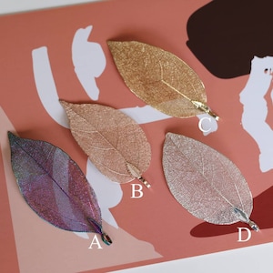 May include: Four delicate leaf-shaped pendants with a metallic finish. The pendants are in shades of gold, pink, silver, and iridescent purple. Each pendant has a small loop at the top for attaching to a chain or cord.