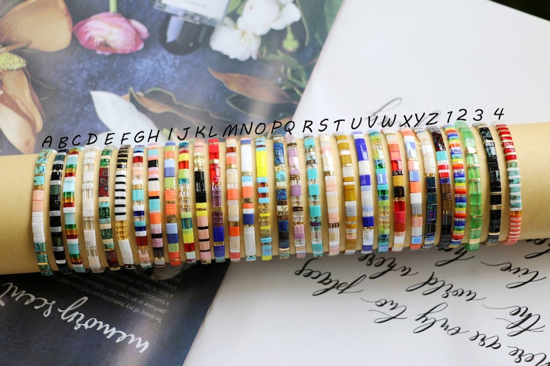 Pu&ograve; includere: Una collezione di braccialetti colorati con perline e una variet&agrave; di motivi. I braccialetti sono impilati uno sopra l'altro ed etichettati con lettere e numeri.