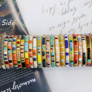 Pu&ograve; includere: Una collezione di braccialetti colorati con perline impilati su una superficie. I braccialetti sono fatti di piccole perline quadrate di vari colori e motivi.