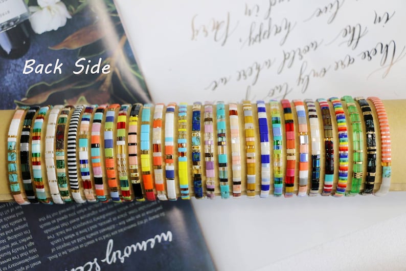 Pu&ograve; includere: Una collezione di braccialetti colorati con perline impilati su una superficie. I braccialetti sono fatti di piccole perline quadrate di vari colori e motivi.