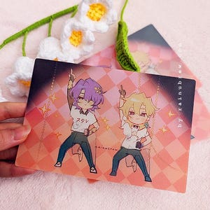 May include: A pink and white illustrated card with two anime characters. The characters are wearing casual clothing and are posing with their arms raised. The text "スノ" is visible on the card.
