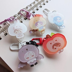 May include: A collection of colorful acrylic keychains featuring cartoon characters. The keychains include a bunny, a bear, a bird, and an apple. Each keychain has a metal clasp for easy attachment. The background includes a spiral notebook.