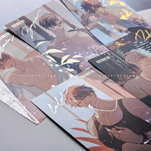 May include: A collection of art prints featuring illustrations of male figures in various poses. Each print has a unique design, with soft color palettes and artistic details. Some prints include handwritten names and website addresses.