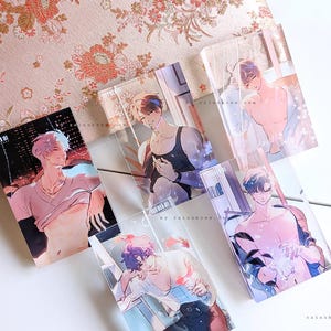 May include: A collection of transparent acrylic art prints featuring male figures in various poses. The prints showcase illustrations with soft color palettes, set against a floral patterned background and a white surface. The art style is reminiscent of anime.