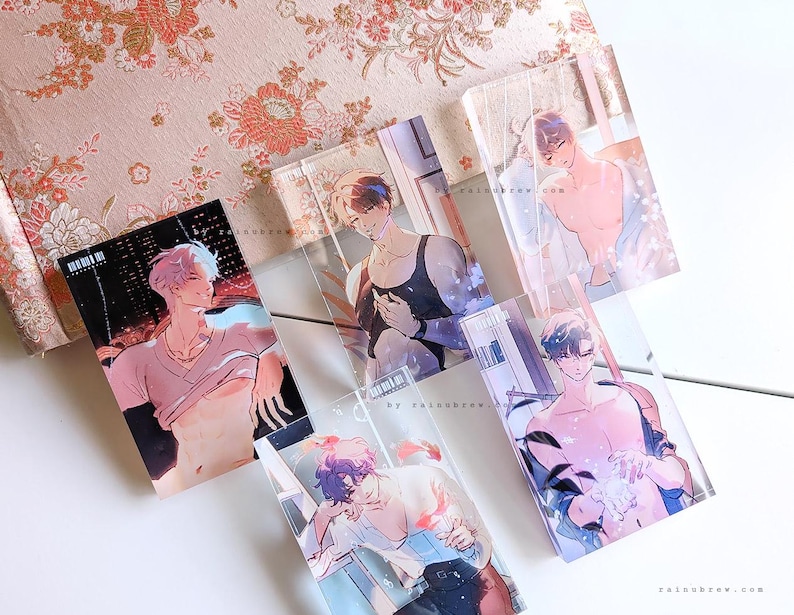 May include: A collection of transparent acrylic art prints featuring male figures in various poses. The prints showcase illustrations with soft color palettes, set against a floral patterned background and a white surface. The art style is reminiscent of anime.