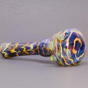 May include: A hand pipe made of glass featuring a deep blue base color with swirling patterns of yellow and orange throughout the stem and bowl. The bowl section includes three small, raised, light green glass dots arranged around the exterior.