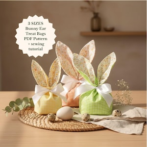 May include: Three fabric bunny ear treat bags in yellow, peach, and green gingham patterns, tied with white ribbons. The bunny ears have floral patterns. The bags are on a woven mat with eggs.