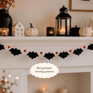 May include: A decorative garland featuring alternating black felt bat shapes and small orange felt balls strung together on a thin cord. The text "Bat garland sewing pattern" is printed in black on a white scalloped oval shape in the centre.