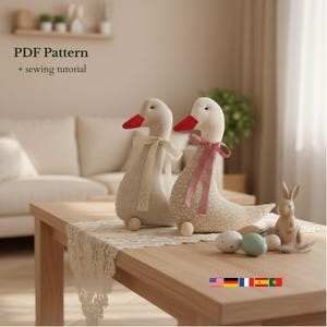 Peut inclure: Deux poupées d'oie en peluche avec des becs rouges et des corps crème, l'une avec un nœud en dentelle et l'autre avec un nœud vichy rouge et blanc. Les poupées sont sur une table en bois avec un chemin de table en dentelle, ainsi qu'un lapin en céramique et des œufs peints. Texte sur l'image : PDF Pattern + sewing tutorial.