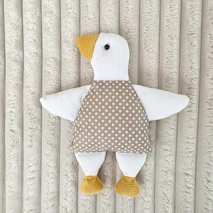 Könnte beinhalten: Ein handgefertigtes weißes Gänse-Plüschtier mit gelbem Schnabel und Füßen. Die Gans trägt ein braunes Kleid mit weißen Punkten. Das Spielzeug hat ausgebreitete Flügel und ein schwarzes Auge.