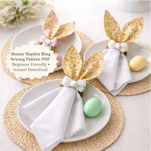 May include: Three white plates with folded white napkins, each adorned with a bunny ear napkin ring. The bunny ears are made of fabric with a floral pattern. The text "Bunny Napkin Ring Sewing Pattern PDF" is visible.