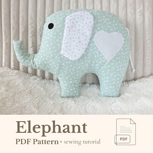 May include: A mint green elephant-shaped pillow with white floral patterns and a white heart applique. The elephant has a black eye and white ears with a floral pattern. The text "Elephant PDF Pattern + sewing tutorial" is below the pillow.