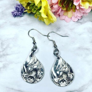 May include: Silver teardrop shaped earrings with a floral design. The earrings have a silver hook closure.