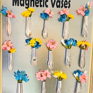 May include: A silver metal frame with the text "Magnetic Vases" at the top.  The frame contains 12 silver spoons with the handles cut off and used as vases.  Each vase holds a small bouquet of flowers in pink, yellow, and blue.