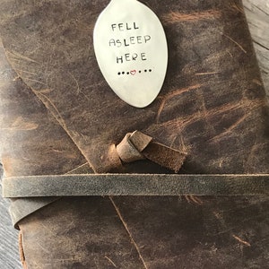 May include: A brown leather journal with a silver spoon bookmark that says "FELL ASLEEP HERE" with two red hearts.