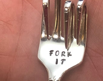 May include: A silver fork bent into the shape of a middle finger with a key ring attached. The fork has the words "Fork It" engraved on it.