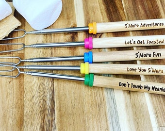 May include: Five wooden roasting sticks with metal prongs and colorful tips. The sticks are engraved with fun sayings: "S'more Adventures", "Let's Get Toasted", "S'more Fun", "Love You S'more", and "Don't Touch My Weenie".