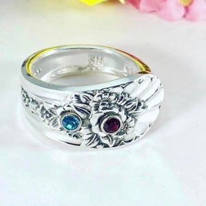 Puede incluir: Un anillo de plata con un diseño floral y dos piedras preciosas. El anillo tiene una piedra azul y una piedra roja.