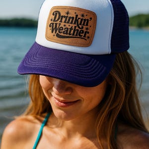 May include: A white and navy blue trucker hat with a purple brim. The hat features a brown leather patch with the words "Drinkin' Weather" in black script and star accents. The hat is worn by a person.