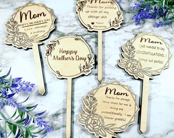 May include: Five wooden signs with floral designs and heartfelt messages for Mother's Day. The signs are attached to wooden sticks, making them ideal for planting in a garden or displaying in a vase.